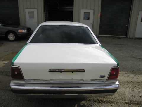 1991 Ford LTD Crown Victoria - Straight-on rear view of a white full-size Ford sedan with green trim and Ford oval, parked on gravel before garage doors.