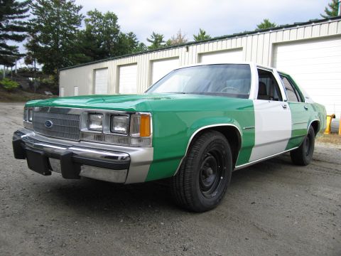 1991 Ford LTD Crown Victoria - two-tone green-and-white Ford sedan with blacked-out steel wheels parked on gravel in front of a metal storage building.