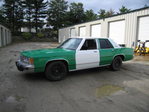 1991 Ford LTD Crown Victoria - two-tone green-and-white Ford sedan with steel wheels parked on a gravel lot beside metal storage buildings.