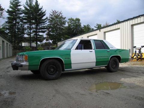 1991 Ford LTD Crown Victoria - two-tone green-and-white Ford sedan viewed from the front-left, parked on gravel near metal garages and pine trees.