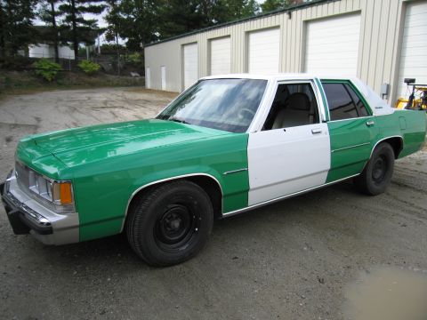 1991 Ford LTD Crown Victoria - two-tone green-and-white four-door sedan with steel wheels parked at an angle on gravel near metal garages.