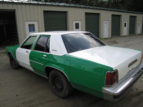 1991 Ford LTD Crown Victoria - green and white two-tone sedan shot from the rear quarter, parked on gravel in front of green storage-unit doors.