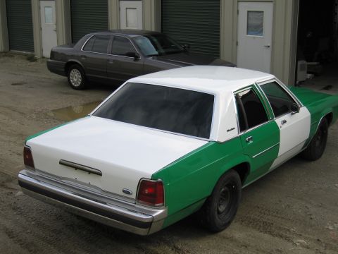 1991 Ford LTD Crown Victoria - white-and-green two-tone Ford sedan seen from the rear, with a gray sedan and storage units in the background.