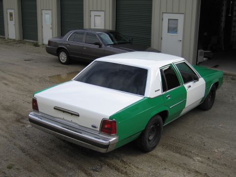 1991 Ford LTD Crown Victoria - White-and-green two-tone Ford sedan from the rear passenger side, parked on gravel near self-storage doors and a gray sedan.