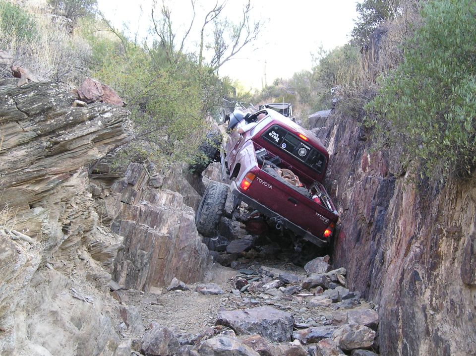 Maroon Toyota pickup truck stuck on a steep, rocky incline. The truck is tilted at a sharp angle