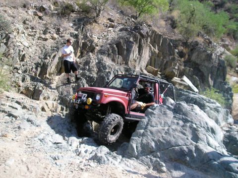 1986 Toyota Hilux - A red off-road vehicle navigates a challenging rocky incline with a person standing on the rocks above.