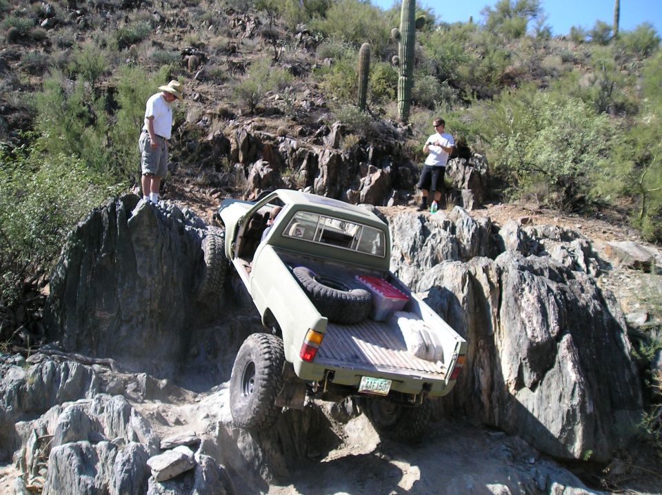 Off-road pickup truck with large tires climbing a rocky incline, spare tire in bed. Two people watch from above