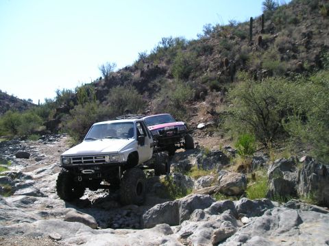 1986 Toyota Hilux - Two modified trucks navigate a challenging rocky path under a clear blue sky.