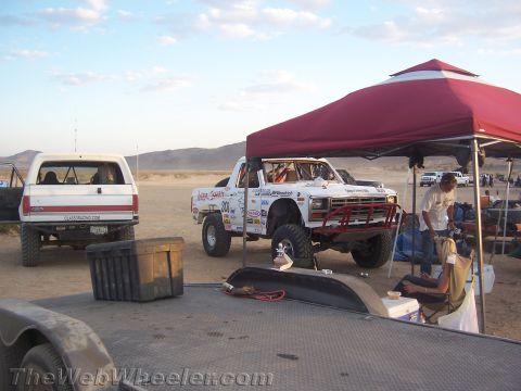 1990 Ford Bronco - Two off-road race trucks sit at a desert pit area, one under a red pop-up canopy as a crew member works nearby.