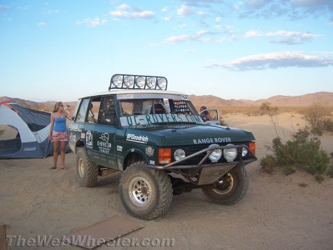 1990 Ford Bronco - green Range Rover prepped for off-road racing with a roof light bar sits in the desert near a tent as a woman stands beside it.