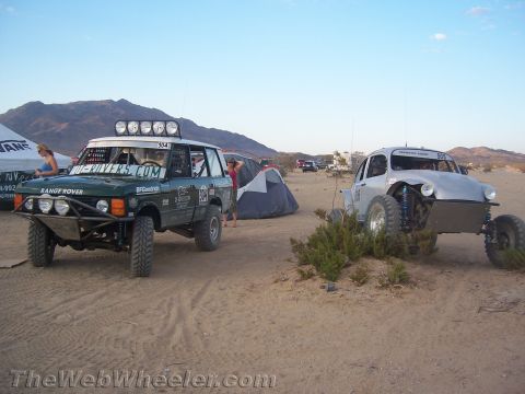 1990 Ford Bronco - green Range Rover off-road racer and a silver Baja Bug rest at a desert camp with tents and mountains behind.