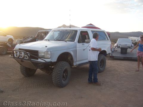 1990 Ford Bronco - white Ford Bronco set up for off-road racing with front light bar sits at a desert camp as people stand alongside.
