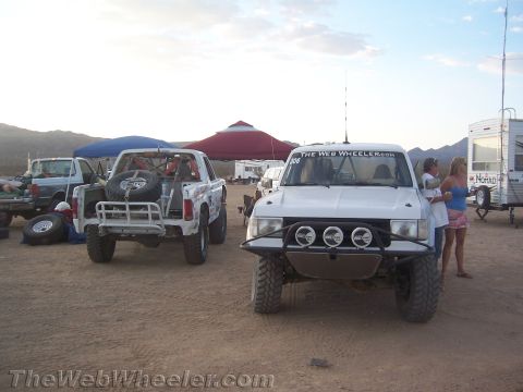 1990 Ford Bronco - Two off-road race trucks, one white with a triple-light front bumper, sit at a dusty desert camp with trailers and canopies.