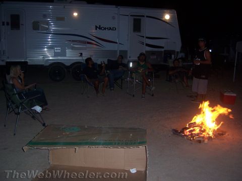 1990 Ford Bronco - group sits in camp chairs around a campfire beside a Nomad travel trailer in the dark desert night.