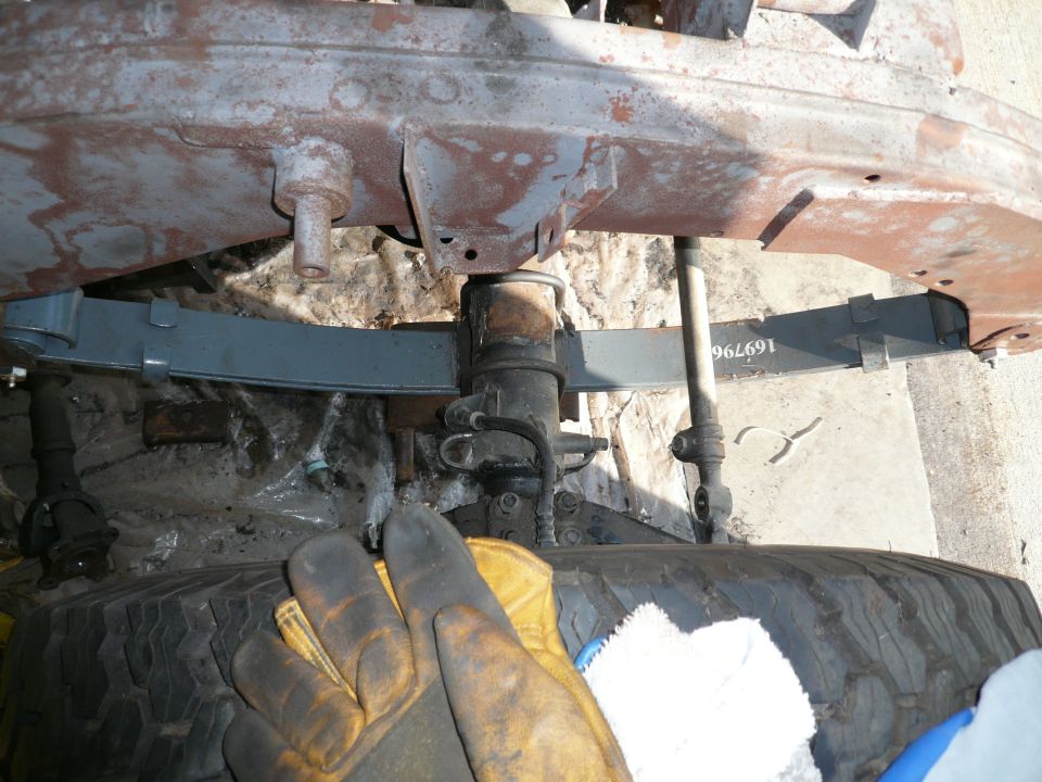Front parabolic leaf spring and suspension under a rusty chassis, with gloved hand and tire