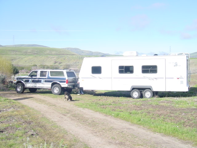 White and blue lifted Ford SUV towing a white travel trailer on a dirt road, dog sitting nearby.