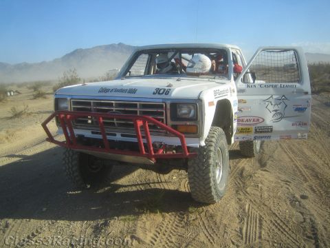 1990 Ford Bronco - white off-road race truck numbered 306 with a red tube bumper and open door sits in the dusty desert with a helmeted driver inside.
