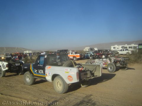 1990 Ford Bronco - Several off-road race trucks, including number 1474, line up on a dusty desert staging area with RVs in the background.