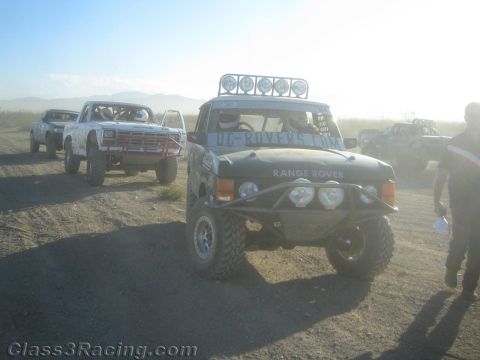 1990 Ford Bronco - green Range Rover off-road racer with a roof light bar leads other race trucks down a dusty desert road.