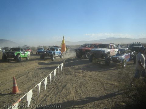 1990 Ford Bronco - lineup of off-road race trucks waits behind a pennant flag line and traffic cone in the dusty desert.