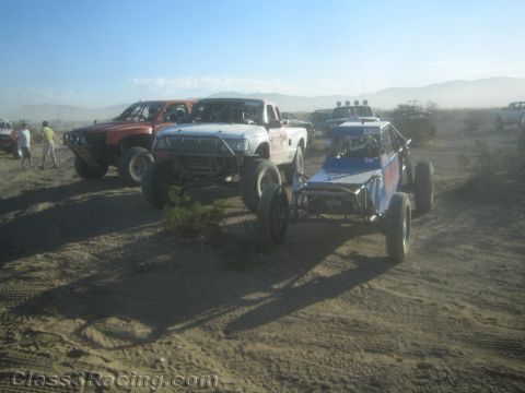 1990 Ford Bronco - white off-road race truck and a blue tube-frame sand buggy sit in a dusty desert staging area with other vehicles.