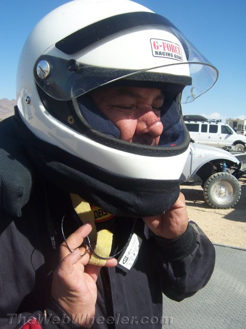1990 Ford Bronco - man fastens the chin strap of his white G-Force racing helmet with the visor up, an off-road vehicle behind him in the desert.