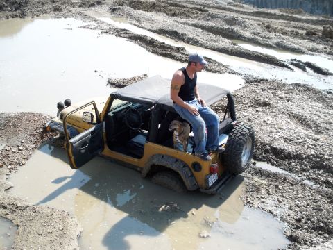 1996 Ford Bronco - A yellow Jeep Wrangler Rubicon is stuck in a muddy pit with a man sitting on the back and a dog inside.
