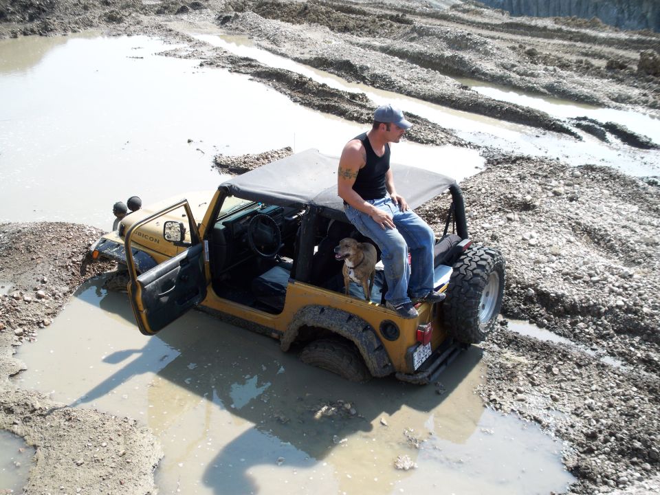 Yellow Jeep Wrangler Rubicon stuck in mud, driver sitting on the back, dog inside