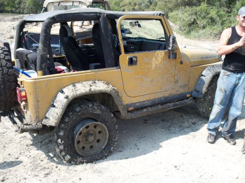 1996 Ford Bronco - A yellow Jeep Wrangler, covered in mud, sits with a flat tire. Another vehicle is visible in the background.