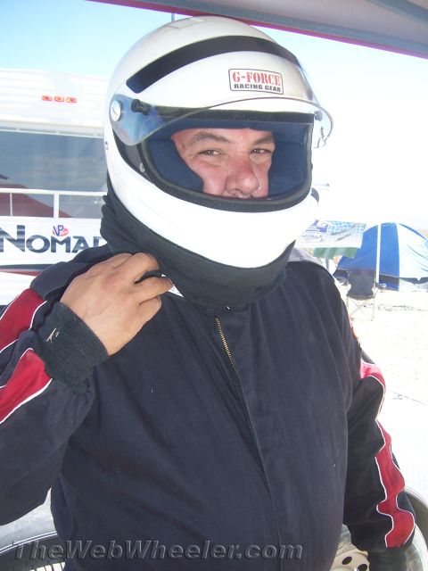 1990 Ford Bronco - man wearing a white G-Force full-face racing helmet and black balaclava under a canopy at a dusty desert venue.