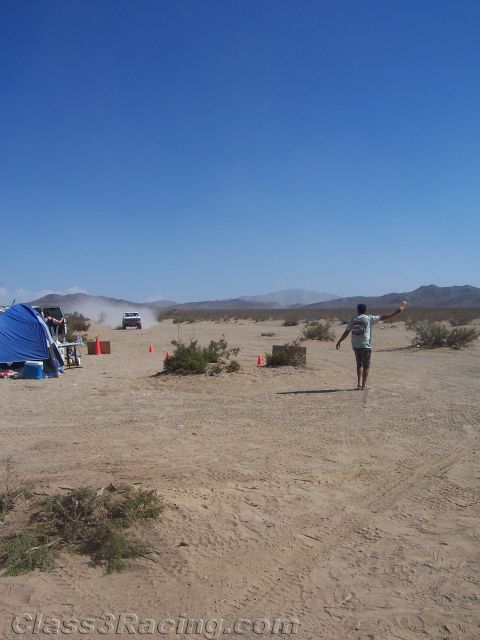 1990 Ford Bronco - person stands in a flat open desert beside camp tents and traffic cones as a distant vehicle kicks up dust under a clear blue sky.