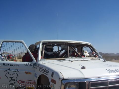 1990 Ford Bronco - white off-road race truck with a roll cage and window nets holds helmeted crew members, marked with sponsor decals in the desert.