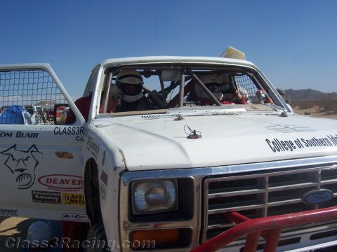 1990 Ford Bronco - front end of a white Ford off-road race truck with a roll cage, window nets, sponsor decals and helmeted occupants in the desert.