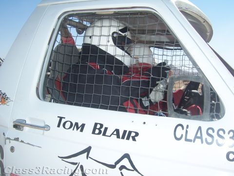 1990 Ford Bronco - Side window-net door of a white off-road race truck lettered Tom Blair, with helmeted, harnessed occupants visible inside.