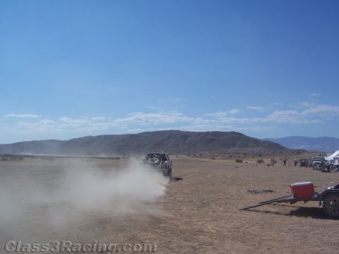 1990 Ford Bronco - off-road vehicle raises a large dust cloud as it drives across a desert flat with mountains and a trailer in the background.