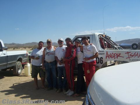 1990 Ford Bronco - group of people gather beside a white off-road race truck lettered Anger Issues, with a pickup and gas cans nearby in the desert.