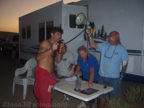 1990 Ford Bronco - shirtless man speaks into a handheld megaphone held by another man at an outdoor folding table beside an RV at dusk.