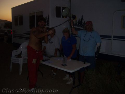 1990 Ford Bronco - dim dusk scene of several people gathered around a white folding table beside an RV trailer in the desert.