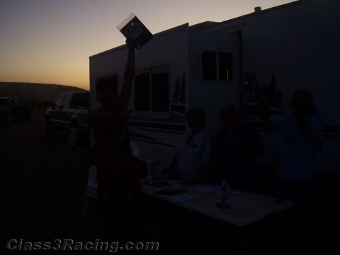 1990 Ford Bronco - silhouetted shirtless man holds up a plaque overhead at dusk beside an RV trailer as others stand at a table.