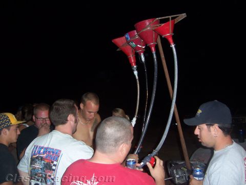 1990 Ford Bronco - nighttime crowd gathers around a wooden stand fitted with red funnels and hoses for drinking, holding cans.
