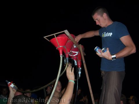 1990 Ford Bronco - man in a blue Hollister shirt stands atop a wooden rig of red funnels and hoses at night, holding beer cans over a crowd.