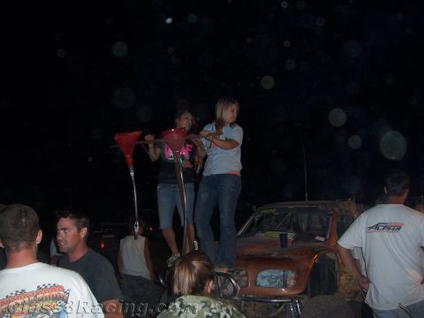 1990 Ford Bronco - Two women stand atop an off-road race truck at night beside a red funnel rig as a crowd of people gathers around.