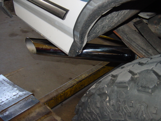 Chrome exhaust tip below white rocker panel and black fender flare, with off-road tire and metal in a shop