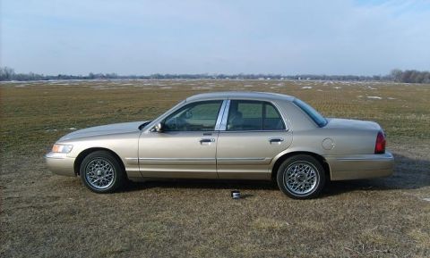 2000 Mercury Grand Marquis - Side view of a gold 2000 Mercury Grand Marquis sedan on a grassy field under a cloudy sky.