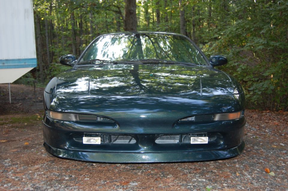 Front view of a dark green coupe with pop-up headlights and fog lights. The car is parked outdoors near trees