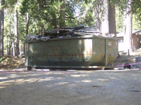 1986 Ford Bronco - A large green dumpster sits on a dirt lot, filled with various pieces of trash and debris.