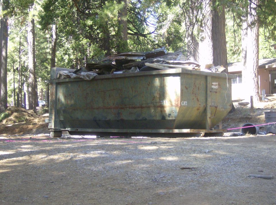 Green dumpster filled with debris, parked on a dirt lot with trees and a building in the background