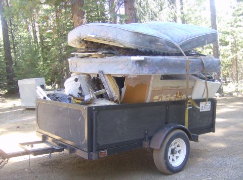 1986 Ford Bronco - Trailer filled with old mattresses and construction waste is parked in a wooded area.