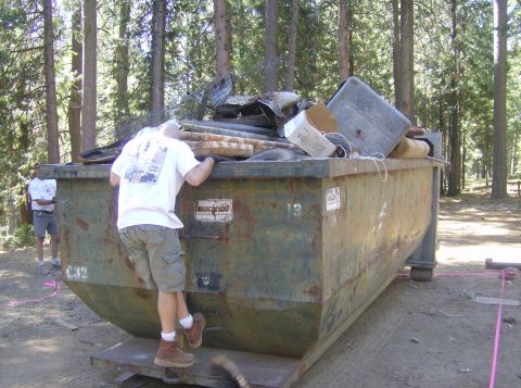 1986 Ford Bronco - A man peers into a large, rusty dumpster overflowing with various discarded items in a forest setting.
