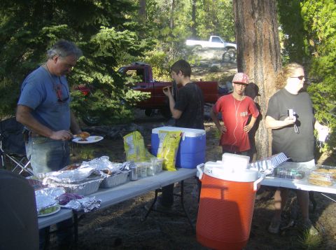 1986 Ford Bronco - A group of people are gathered around picnic tables with food, with a red pickup truck parked behind them and a white pickup truck visible…